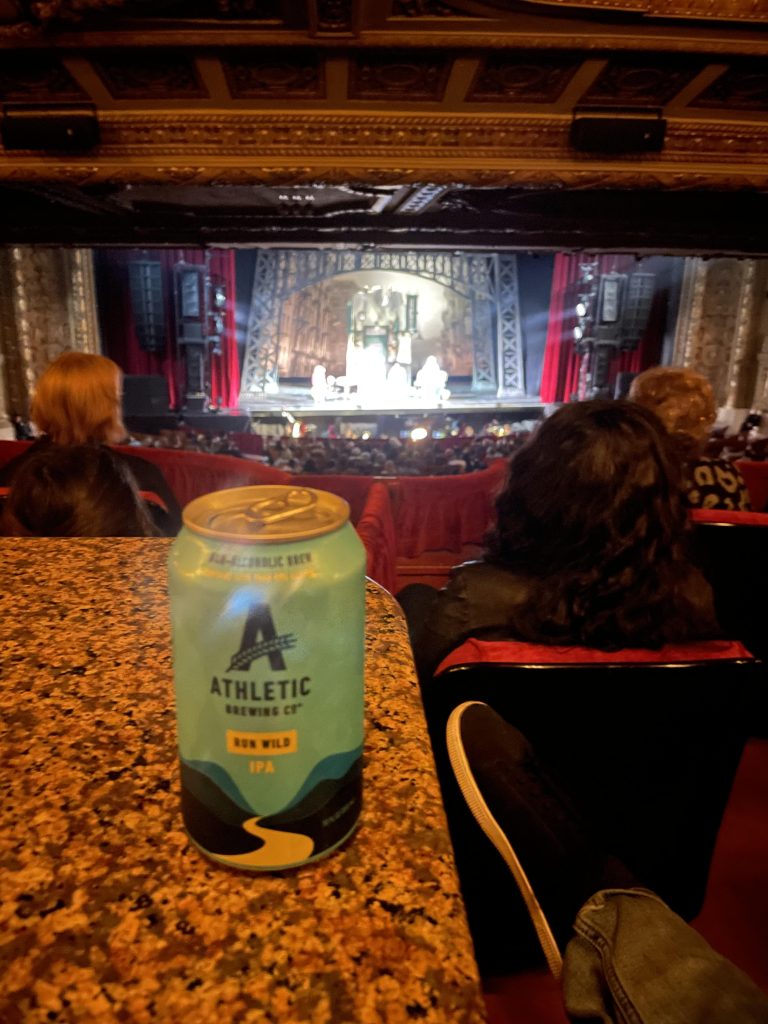 Can of Athletic Brewing Company Run Wild IPA non-alcoholic beer resting on a balcony railing at the Chicago Theatre. The ornate interior and stage are visible in the background, with an audience seated below enjoying the performance.