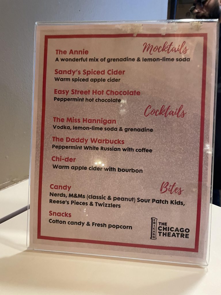 Photo of the mocktail menu at the Chicago Theatre, taken in November 2024. The menu features mocktails like 'The Annie' (grenadine and lemon-lime soda), 'Sandy’s Spiced Cider' (warm spiced apple cider), and 'Easy Street Hot Chocolate' (peppermint hot chocolate). It also lists cocktails, candy options like M&Ms and Sour Patch Kids, and snacks such as cotton candy and fresh popcorn.